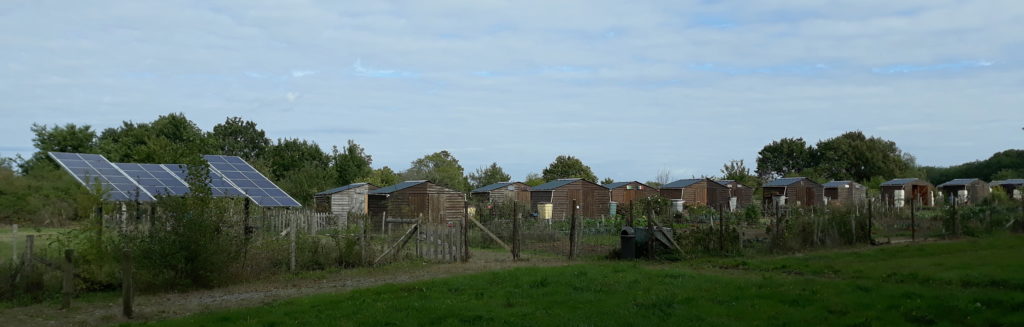 Vue des jardins partagés de Donges : panneaux solaires photovoltaïques au sol, série de cabanes en bois avec récupérateur d'eau et parcelles individuelles, le tout enclos d'une clôture