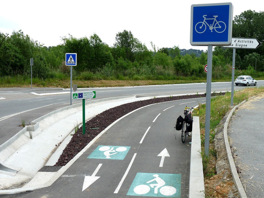 Piste cyclable en campagne, sécurisée, séparée de la route par un parapet en béton. Piste neuve, en bon état, peinture propre au sol, panneaux routiers spécifiques vélo