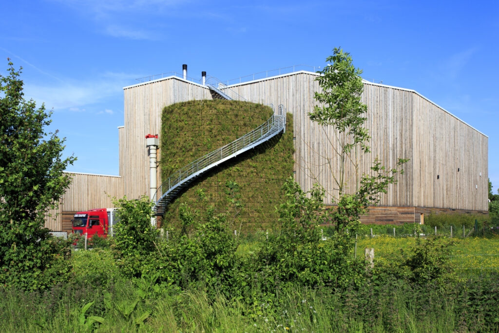 Mur végétalisé d'une entreprise : vue d'un réservoir industriel entièrement recouverte de végétation. Haie en premier plan et bâtiment industriel recouvert de bardage bois en arrière-plan